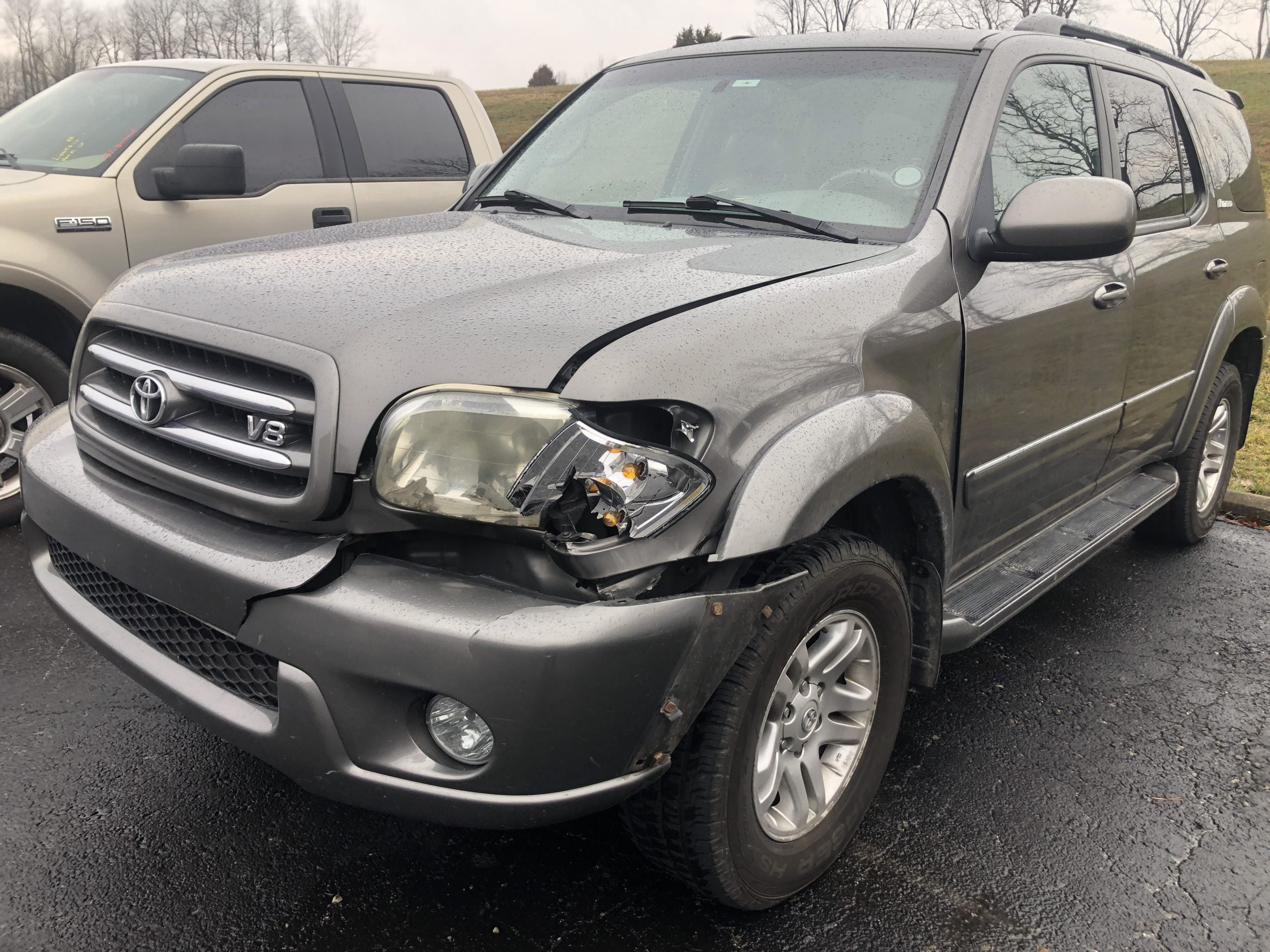 A gray Toyota SUV with a damaged front end, including a broken right headlight and dented bumper, parked on a wet asphalt surface beside another vehicle on a cloudy day.