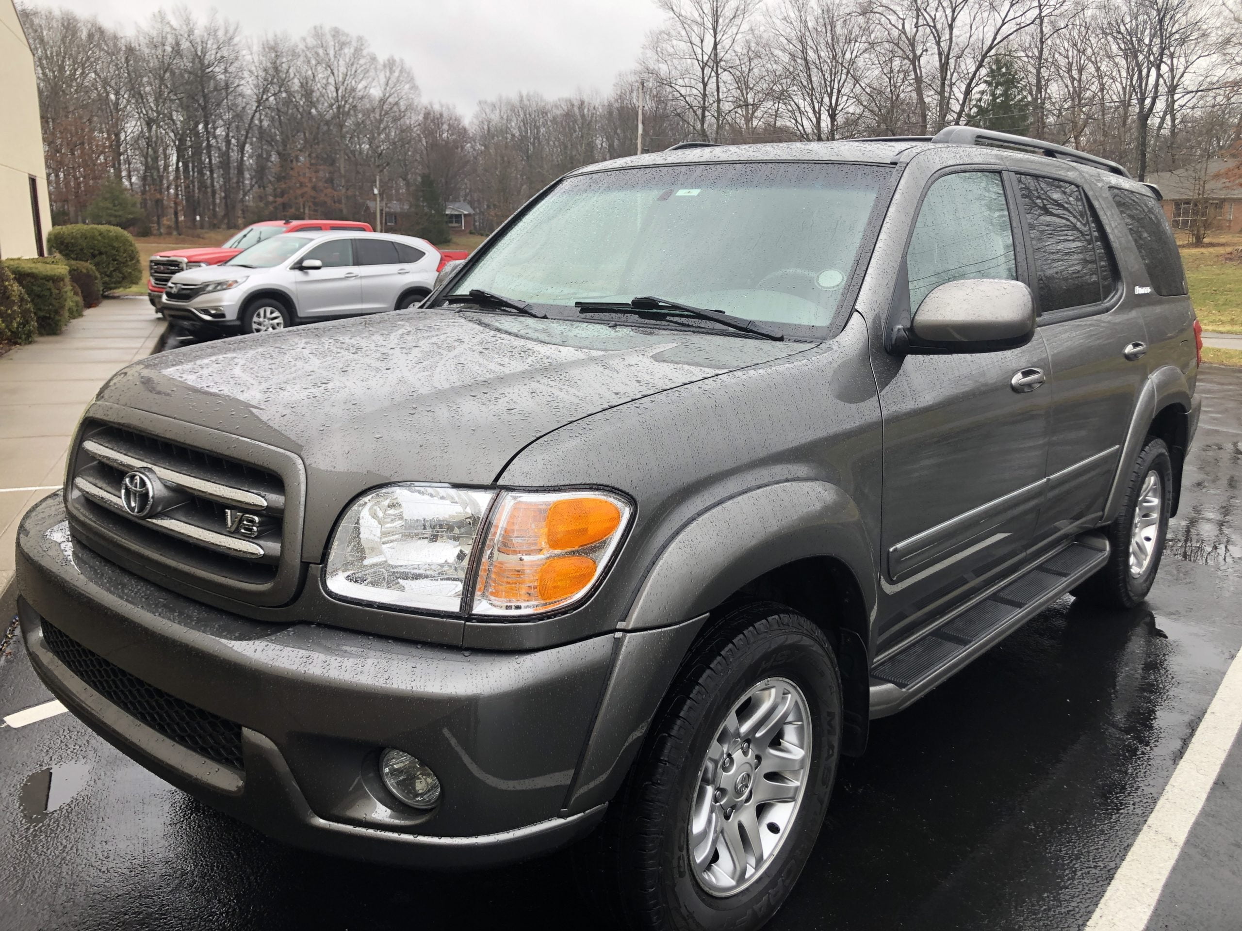 A gray Toyota Sequoia SUV parked in a lot on a rainy day, with water droplets visible on its surface and other vehicles and bare trees in the background.