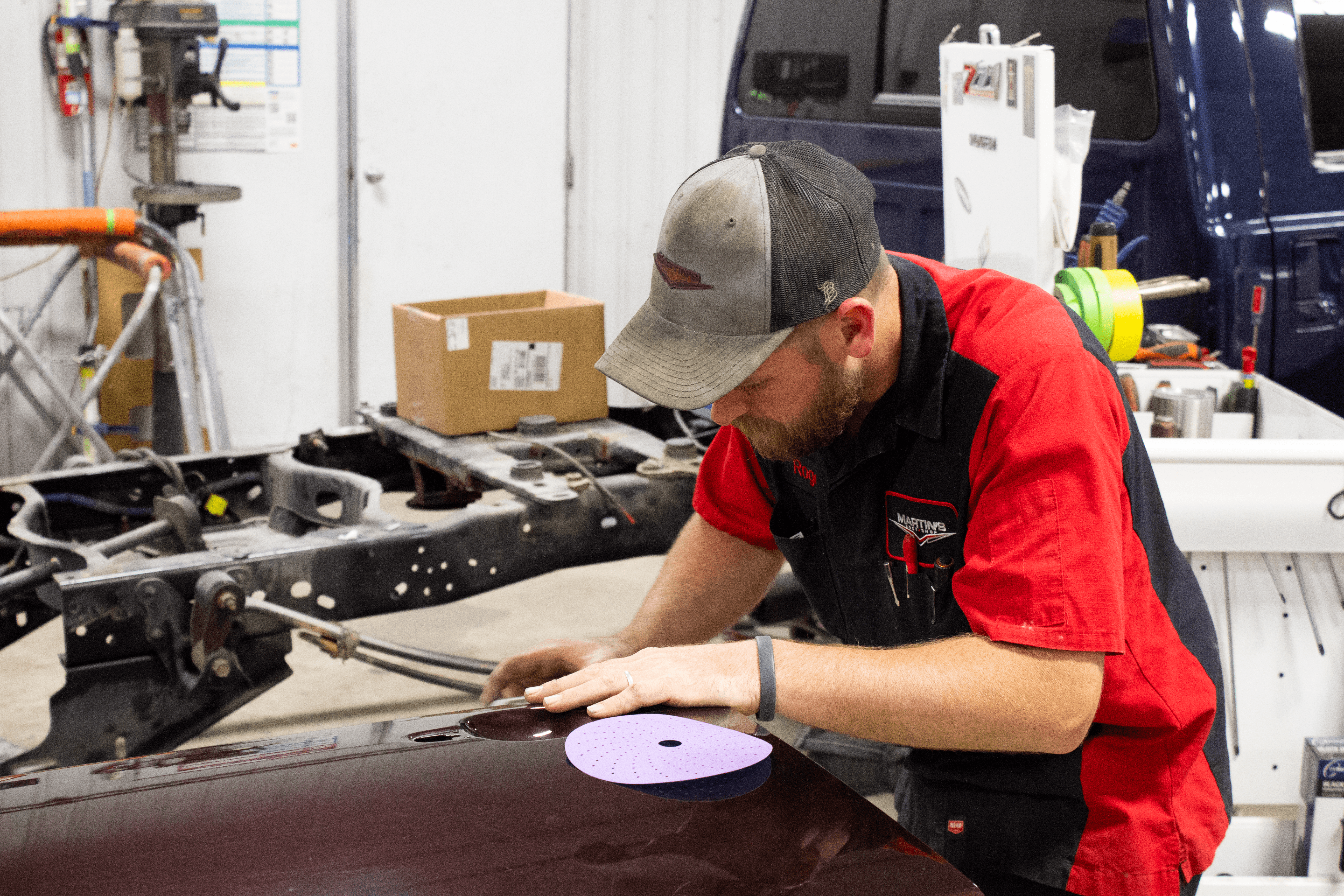 IMG_4921 A member of the Martin's Body Shop team in a red and black uniform carefully sands the surface of a maroon car hood in an auto repair shop, with tools, a blue truck, and equipment visible in the background.