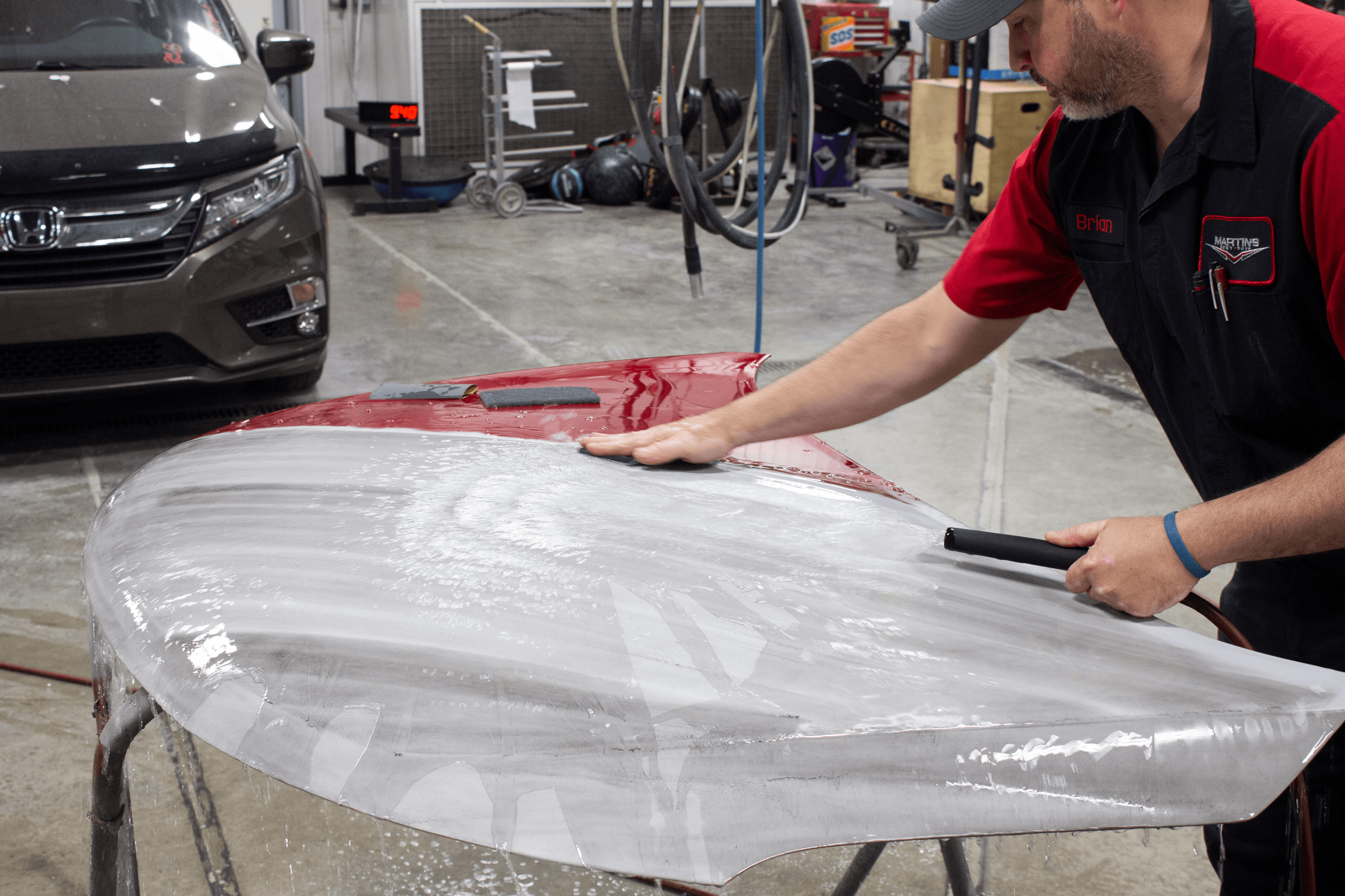 IMG_4924 A man wearing a work uniform cleans and sands the surface of a car hood in Martin's Body Shop. A brown car, workshop tools, and various garage items are visible in the background as part of the dedicated Martin's Body Shop team.