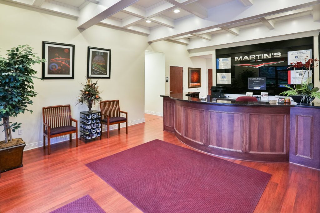 martinsbodyshop-1 Reception area with a curved wooden desk, computer monitors, maroon rugs, two chairs, wall art, a potted plant, and a flower arrangement. Signs reading MARTIN’S highlight the auto body repair steps in this well-lit and tidy space.