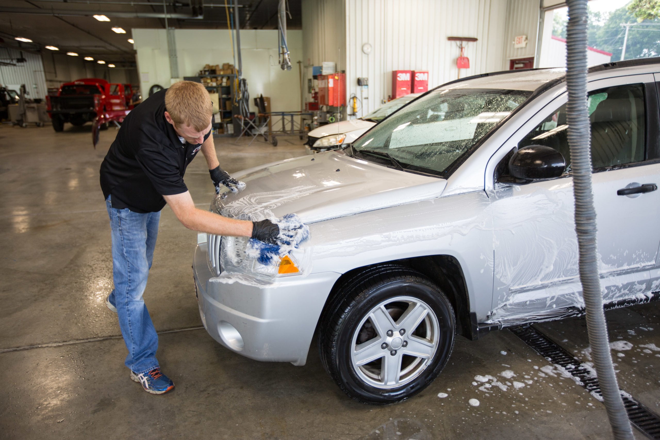 A person wearing gloves washes the hood of a silver SUV with a soapy sponge inside a spacious, well-lit automotive garage, an essential part of the auto body repair steps. Cleaning equipment is visible in the background.