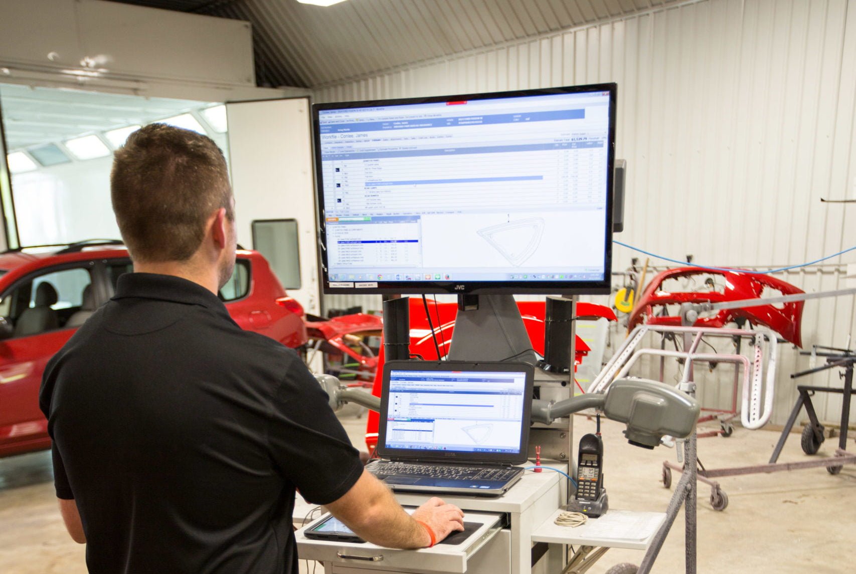 A person in a black shirt uses a computer workstation with a large monitor in an auto repair shop, reviewing auto body repair steps, with car parts and a red vehicle visible in the background.