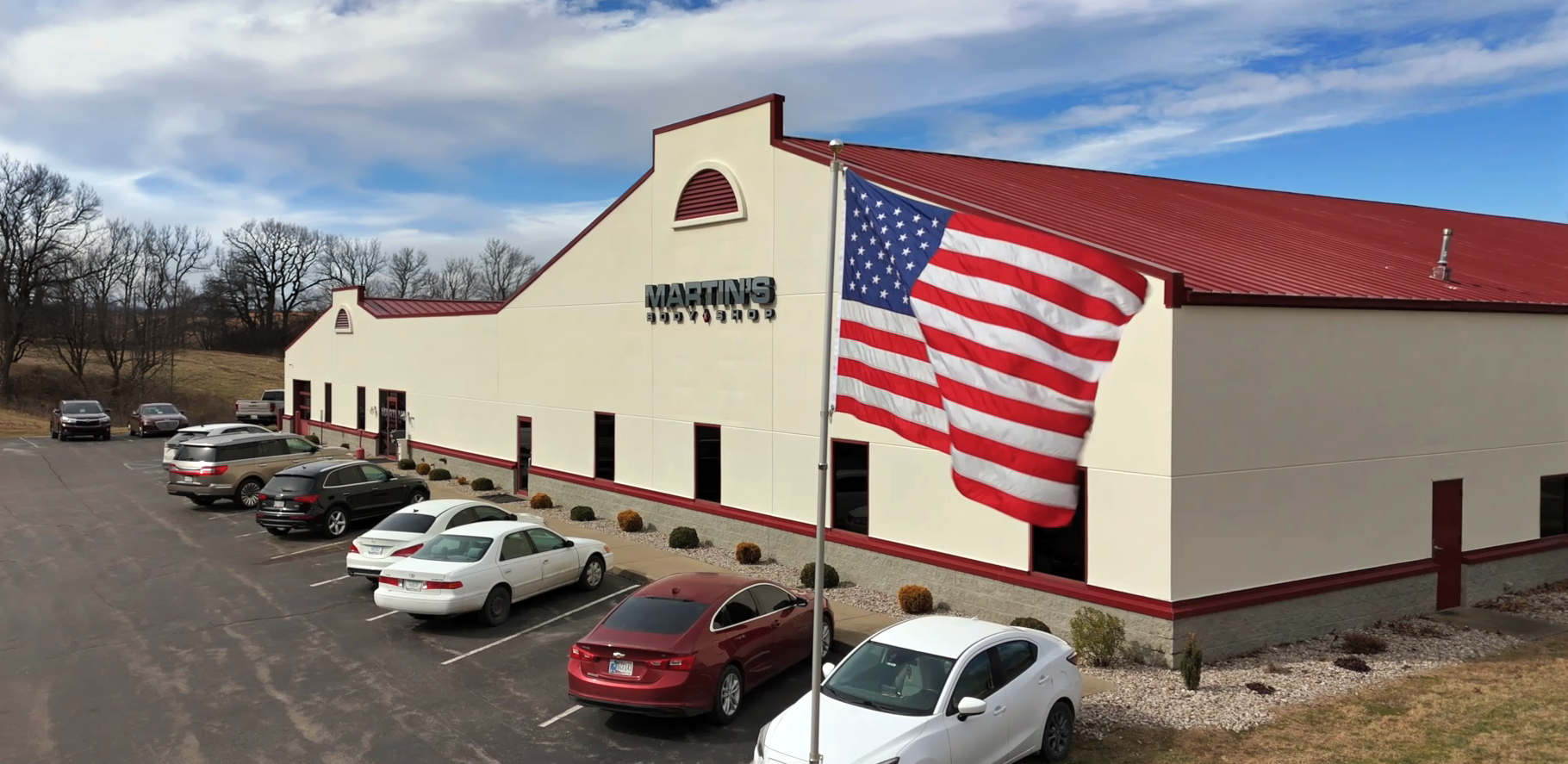 NewSalisbury_Drone_Edit A large beige building with a red roof labeled MARTINS stands behind a parking lot with several cars. An American flag waves on a flagpole in the foreground, and trees and cloudy sky are visible in the background.