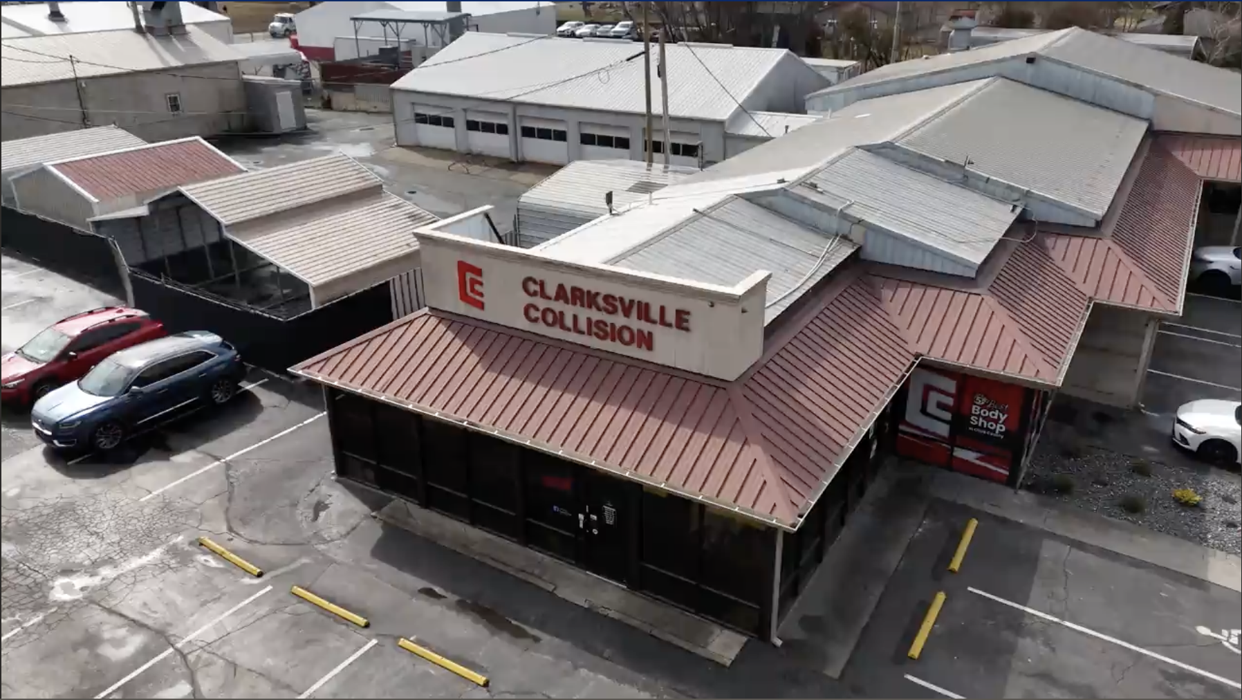Screenshot 2026-05-07 at 11.27.47 AM Aerial view of the Clarksville Collision auto body shop, a red-roofed building with a large sign, several parked cars, and an adjacent lot. One of the Martin’s Body Shop locations is visible nearby with additional garages and vehicles.