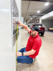 A man in a red shirt and black cap kneels on the floor, measuring the distance from the ground to a checkered calibration chart on a wall inside a spacious garage with cars in the background.