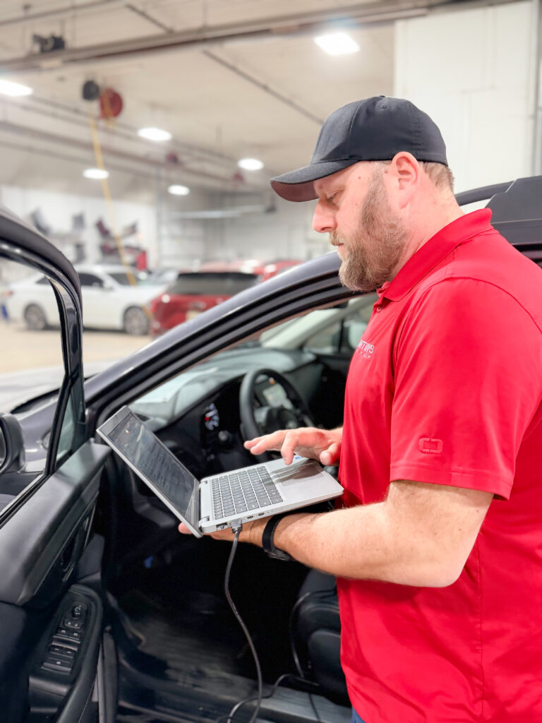 ADAS_2 A man in a red shirt and black cap stands next to an open car door, using a laptop connected to the vehicle inside a well-lit garage with several cars in the background.