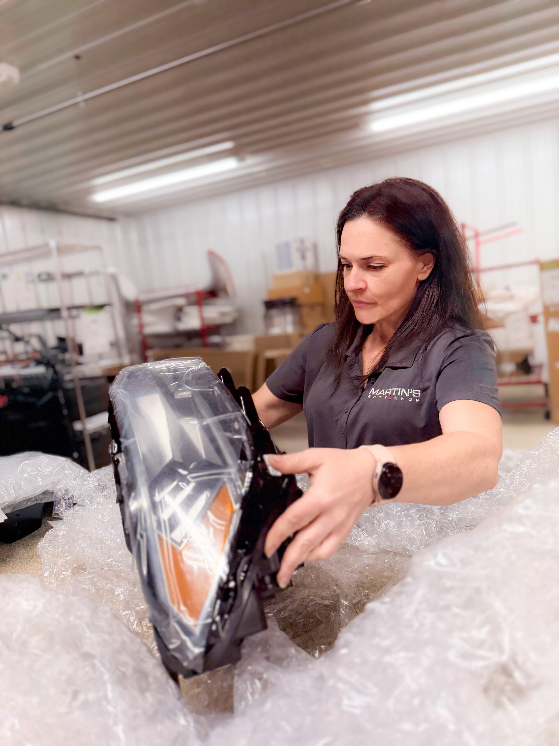 A woman wearing a gray polo shirt opens bubble wrap around a large car headlight inside a workshop with shelves and various equipment, where accurate car repair estimates are essential.