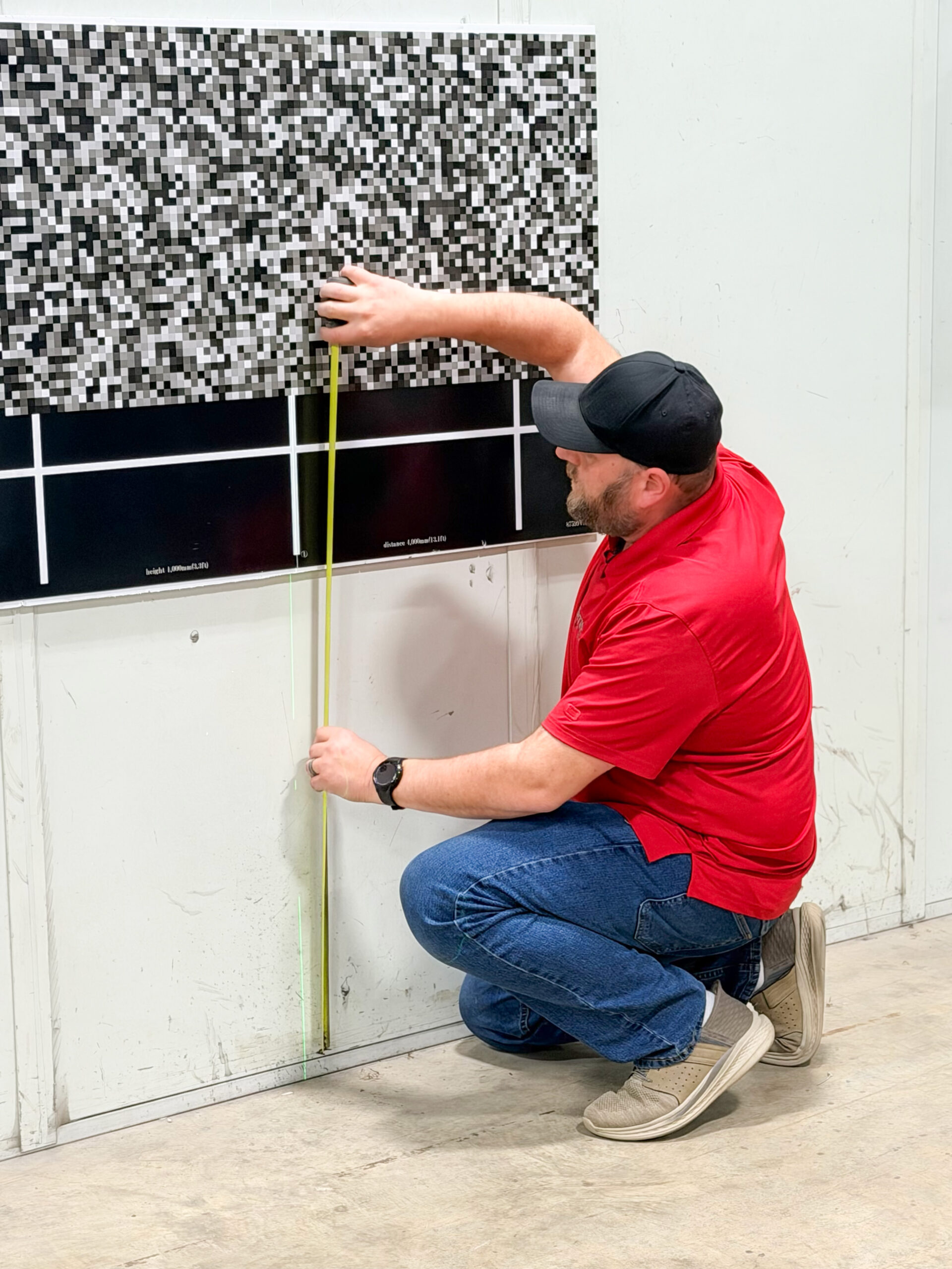 A man in a red shirt and black cap kneels on the floor, measuring the distance from the ground to a large black-and-white pixelated chart on a white wall with a yellow measuring tape, ensuring accurate car repair estimates.