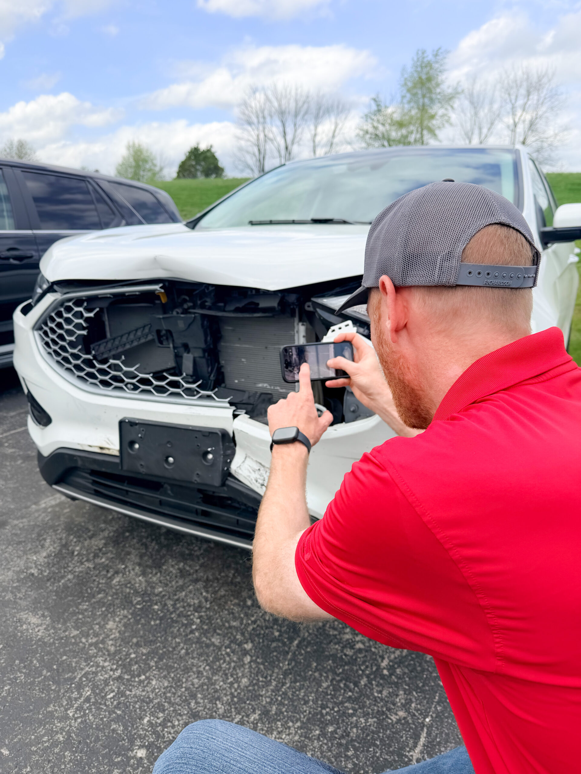 A man in a red shirt and cap takes a photo of the front-end damage on a white SUV with his smartphone in a parking lot, documenting the broken grille and bumper for accurate car repair estimates.