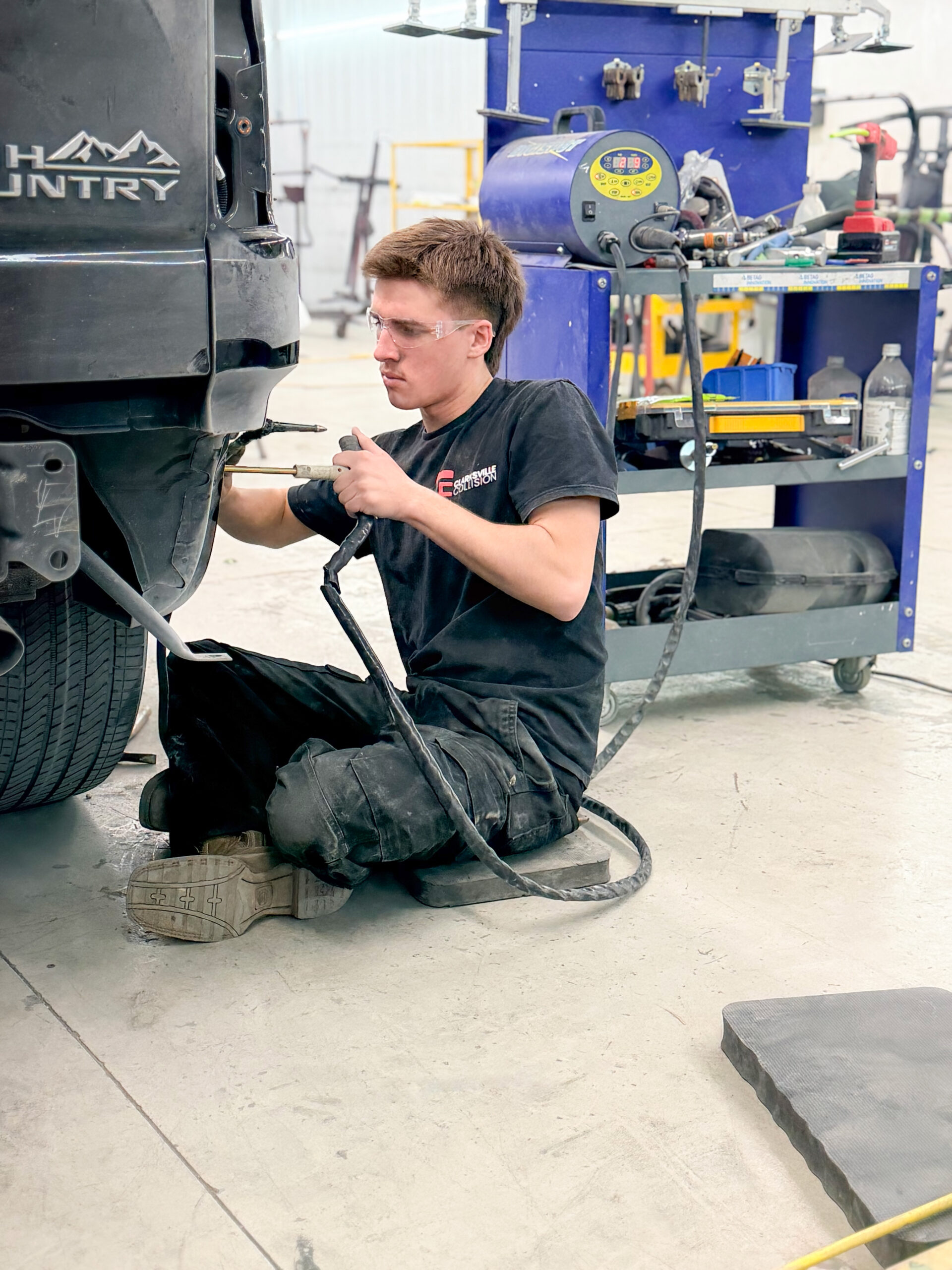 A mechanic in black work clothes kneels on the floor, using a power tool to repair the rear end of a black pickup truck in an auto repair shop known for providing accurate car repair estimates. Tools and equipment are visible in the background.